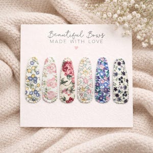 May include: Six fabric hair clips with floral patterns in various colors, including blue, pink, and white, are displayed on a white card. The card reads "Beautiful Bows MADE WITH LOVE". The clips are arranged on a cream-colored surface.