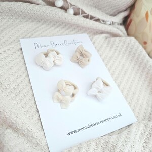 May include: A white card displays four baby headbands with bow accents in shades of cream and beige. The card reads "Mama Bears Creations" and "www.mamabearscreations.co.uk". The headbands are arranged on a textured, off-white surface.