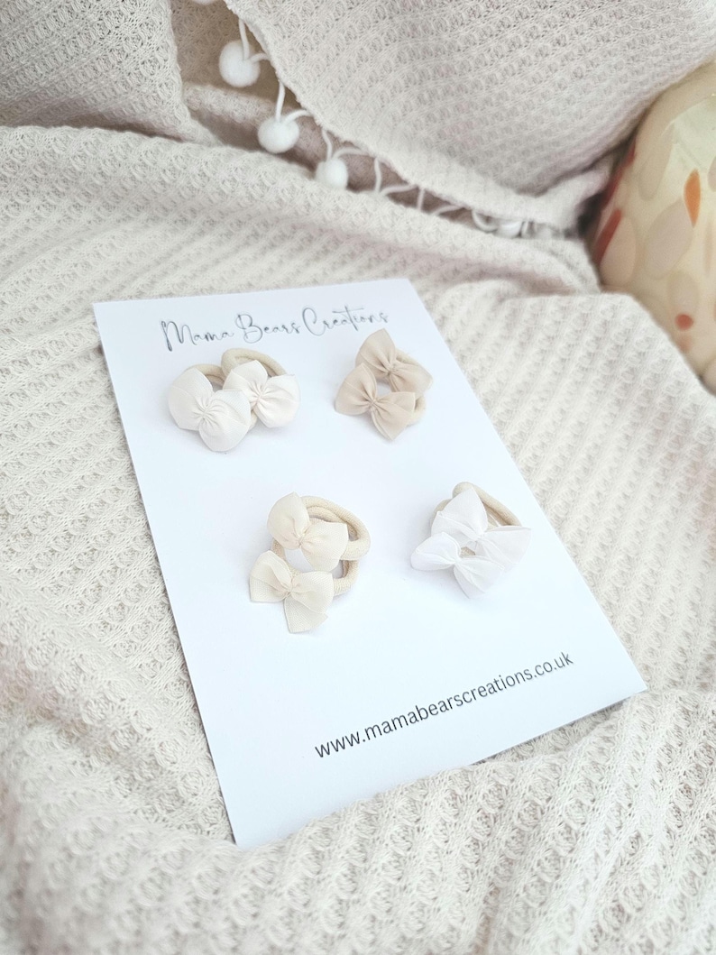 May include: A white card displays four baby headbands with bow accents in shades of cream and beige. The card reads "Mama Bears Creations" and "www.mamabearscreations.co.uk". The headbands are arranged on a textured, off-white surface.