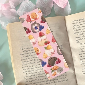 May include: A pink bookmark featuring illustrations of hamsters with various fruits like watermelon, strawberries, and blueberries. The bookmark is rectangular and is placed on an open book.