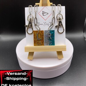 Könnte beinhalten: Zwei mit Glitzer besetzte Buchstaben-Anhänger, einer goldfarben mit dem Buchstaben "L" und einer silberfarben mit dem Buchstaben "R", an silbernen Ketten mit Karabinerhaken. Die Anhänger werden auf einem kleinen Holzständer mit weißem Hintergrund präsentiert.