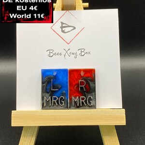 Könnte beinhalten: Zwei kleine, quadratische Harzohrringe mit einem blauen und schwarzen Wirbeldesign auf der linken Seite und einem roten und schwarzen Wirbeldesign auf der rechten Seite. Die Ohrringe sind mit "L MRG" und "R MRG" beschriftet.
