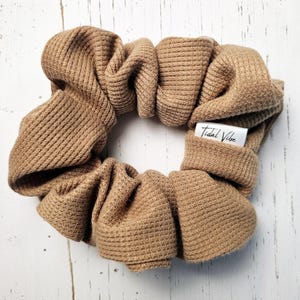 May include: A tan waffle-knit scrunchie with the text "Tidal Vibe" on a small white tag. The scrunchie is made of a soft, textured fabric and is designed to hold hair in place. The scrunchie is a hair accessory.