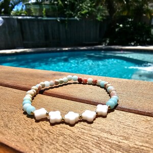 May include: A light blue and pink beaded bracelet with white star-shaped charms. The bracelet is on a wooden surface near a swimming pool.