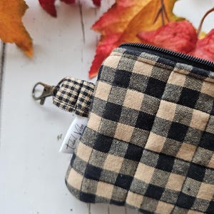May include: A small, beige and black checked pouch with a matching wrist strap and a metal clasp. The pouch has a black zip and a small white tag with text. Autumn leaves in shades of red and orange are in the background.