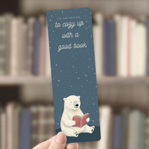 May include: A bookmark featuring a polar bear reading a book. The bookmark has a blue background with white stars and the text "Tis the season to cozy up with a good book."