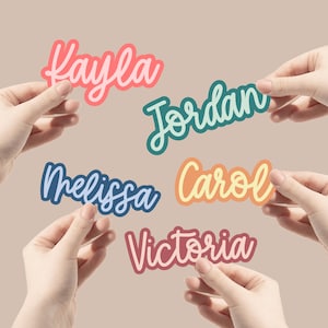 May include: Five colourful name stickers with names Kayla, Jordan, Melissa, Carol, and Victoria. The stickers are held by hands against a light brown background.