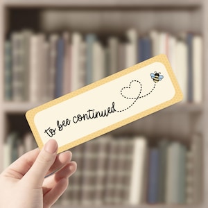 May include: A rectangular bookmark with a pale yellow background and a honeycomb border. The text "to bee continued" is written in black script. A dotted line leads from a heart to a cartoon bee.