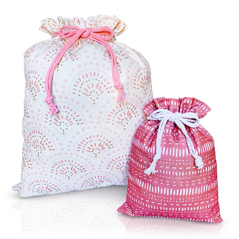 Puede incluir: Dos bolsas de tela rosa con cord&oacute;n de ajuste y diferentes patrones. La bolsa m&aacute;s grande tiene un fondo blanco con dise&ntilde;os geom&eacute;tricos rosa y dorado. La bolsa m&aacute;s peque&ntilde;a tiene un fondo rosa s&oacute;lido con dise&ntilde;os geom&eacute;tricos blancos.