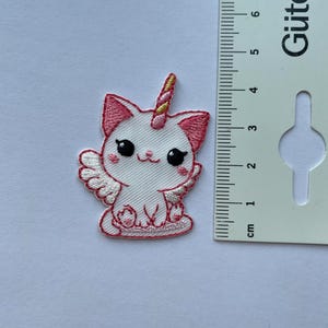 Könnte beinhalten: Ein weiß-rosa bestickter Aufnäher mit einer Katze mit Einhornhorn und Flügeln. Der Aufnäher hat schwarze Augen und einen rosa Umriss. Der Aufnäher ist etwa 5 cm hoch.
