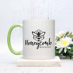 May include: White ceramic mug with a green rim and handle. The mug has a black bee and honeycomb design with the text "Honeycomb Mockups". The mug is sitting on a white marble coaster.