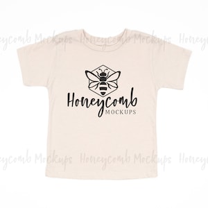Op de afbeelding: Een beige baby-T-shirt met een zwarte bij en honingraat-afbeelding en de tekst "Honeycomb Mockups".