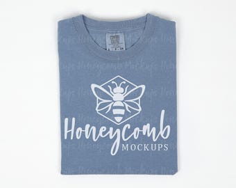 May include: A blue short-sleeved t-shirt featuring a white graphic of a bee inside a hexagon above the text "Honeycomb MOCKUPS". The shirt has a Comfort Colors brand label at the inner neckline, indicating a size small, and a crew neckline.