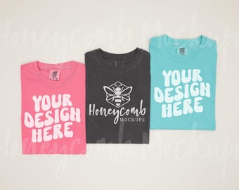 May include: Three short-sleeve t-shirts in pink, charcoal gray, and light blue. The pink and light blue shirts feature the white text 'YOUR DESIGN HERE'. The charcoal gray shirt displays a white bee icon above the text 'Honeycomb MOCKUPS'.