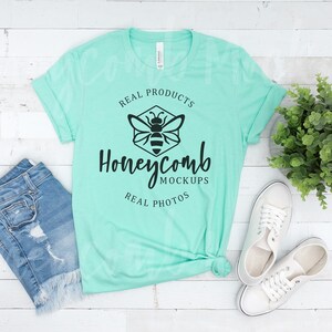 May include: A light turquoise t-shirt with a black bee graphic and the text "Real Products Honeycomb Mockups Real Photos". The t-shirt is laid flat on a white wooden surface with a pair of white canvas shoes and a pair of blue denim shorts.