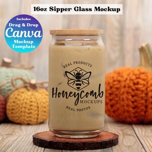 May include: A 16 ounce clear glass sipper with a bamboo lid and a straw. The glass is filled with a light brown liquid and has a black and white logo that says "Honeycomb Real Products" with a bee illustration. The glass is sitting on a wooden coaster in front of a background of knitted pumpkins.