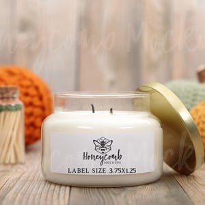 Peut inclure: Une bougie blanche dans un pot en verre transparent avec un couvercle doré. L'étiquette présente une icône d'abeille et le texte "Honeycomb Mockups" avec "LABEL SIZE 3.75X1.25" imprimé en dessous. La bougie a deux mèches.