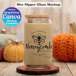 Puede incluir: Un vaso de cristal transparente de 16 oz con tapa de bambú, lleno de una bebida helada de color canela. El vaso presenta un gráfico negro de una abeja dentro de un hexágono con el texto "REAL PRODUCTS Honeycomb MOCKUPS REAL PHOTOS". Una insignia azul dice "Includes Drag & Drop Canva Mockup Template".