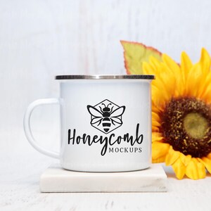 May include: White enamel camping mug with a black bee and honeycomb design and the text "Honeycomb Mockups". The mug is sitting on a white marble coaster and a sunflower is in the background.