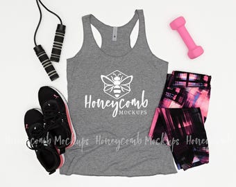 May include: A heather gray racerback tank top featuring a white graphic of a bee inside a hexagon with the text Honeycomb MOCKUPS. The tank is surrounded by a black jump rope, a pink dumbbell, and black and pink patterned athletic leggings.