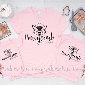 May include: Three pink t-shirts with a black bee logo and the text "Honeycomb Mockups". The shirts are arranged on a white wooden surface with holiday decorations.