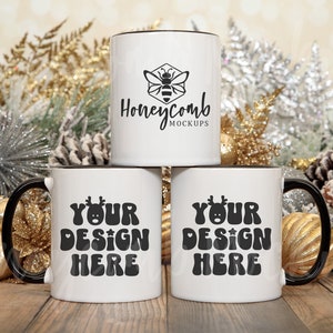 May include: Three white ceramic mugs with black rims and handles are stacked. The top mug has a honeycomb logo with the text "Honeycomb Mockups". The bottom mugs have the text "YOUR DESIGN HERE" in a retro font. The background features holiday decorations.