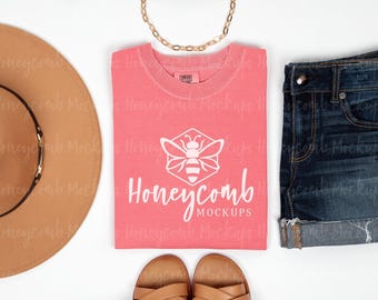 May include: A coral-colored short-sleeve t-shirt featuring a white graphic of a bee inside a hexagon above the text Honeycomb MOCKUPS. The shirt is displayed with a tan wide-brim hat, dark blue denim shorts, and tan sandals.