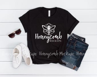 May include: A black t-shirt featuring a white graphic of a bee inside a hexagon with the text Honeycomb MOCKUPS below it. The shirt is displayed alongside a pair of dark blue denim shorts, white sneakers, and brown sunglasses.