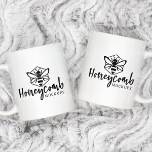 May include: Two white ceramic mugs with black bee and honeycomb logo and the text "Honeycomb MOCKUPS" on a white fluffy background.