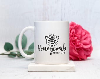Könnte beinhalten: Ein weißer Keramikbecher mit einer schwarzen Grafik einer Biene in einem Sechseck über dem Text Honeycomb MOCKUPS. Der Becher hat einen geschwungenen Henkel und steht auf einem quadratischen weißen Sockel neben einer rosa Blume.