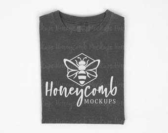 May include: A charcoal gray t-shirt featuring a white graphic of a bee centered inside a hexagon. Below the bee, the text reads Honeycomb MOCKUPS in a white script and sans-serif font. The fabric has a visible texture and a crew neckline.