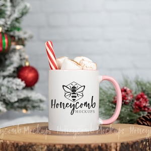 May include: White ceramic mug with a pink handle, filled with hot chocolate and marshmallows. A candy cane sticks out of the top. The mug has a black bee logo and the text "Honeycomb Mockups". The mug is sitting on a wooden slice.