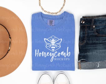 May include: A periwinkle blue crew neck t-shirt with a white graphic of a bee inside a hexagon and the text Honeycomb MOCKUPS, paired with a tan wide-brim hat, dark blue denim shorts, a gold chain necklace, and white sneakers.