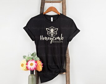 Puede incluir: Una camiseta negra de manga corta con un gráfico blanco de una abeja dentro de un hexágono sobre el texto Honeycomb MOCKUPS. La prenda cuelga de una percha de madera sobre un fondo claro con un jarrón de flores coloridas.