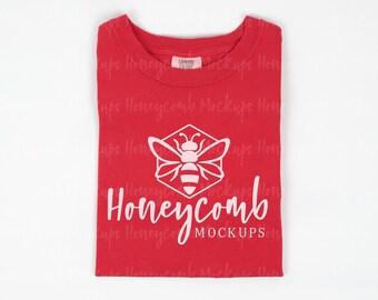 May include: A red short-sleeved t-shirt featuring a white graphic of a bee centered inside a hexagon. Below the bee, the word Honeycomb is printed in a white script font, with the word MOCKUPS in white capital letters underneath.