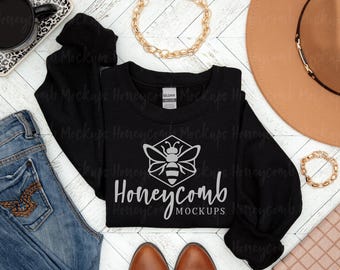 May include: A black long-sleeve crewneck sweatshirt featuring a white graphic of a bee inside a hexagon above the text Honeycomb MOCKUPS. The garment is displayed alongside blue denim jeans, a tan hat, and gold chain jewelry.