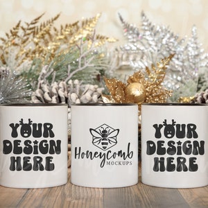 Puede incluir: Tres tazas de cerámica blanca con bordes y asas negras. Las tazas muestran el texto "YOUR DESIGN HERE" en una fuente retro, y una taza presenta un logotipo de abeja con el texto "Honeycomb Mockups". El fondo incluye decoraciones festivas.