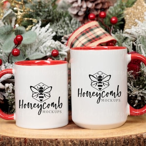 Puede incluir: Dos tazas de cerámica blanca con bordes y asas rojos. Ambas tazas presentan un logotipo de abeja negro con el texto "Honeycomb MOCKUPS". Las tazas están sobre una superficie de madera con un fondo navideño festivo.
