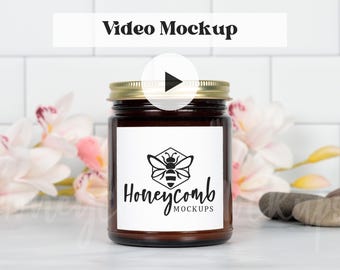 May include: A brown glass candle jar with a gold metal lid and a white rectangular label featuring a black bee icon inside a hexagon and the text Honeycomb MOCKUPS. A play button icon is centered over the top of the jar.