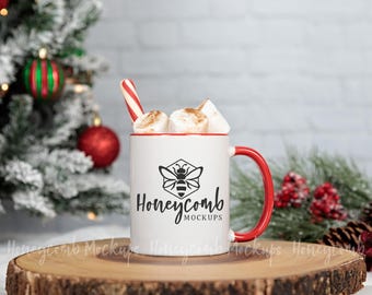 May include: A white ceramic mug with a red rim and handle, featuring a black bee graphic inside a hexagon and the text Honeycomb MOCKUPS. The mug is filled with marshmallows and a red and white striped candy cane.