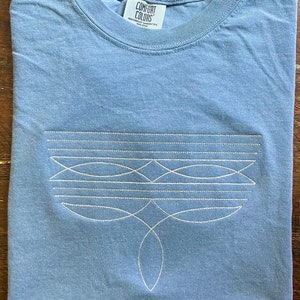 Puede incluir: Una camiseta azul claro con un dise&ntilde;o bordado blanco en la parte delantera. El dise&ntilde;o presenta una serie de l&iacute;neas y formas curvas. La camiseta tiene una etiqueta que dice "Comfort Colors" y "Heavyweight Garment Dyed".