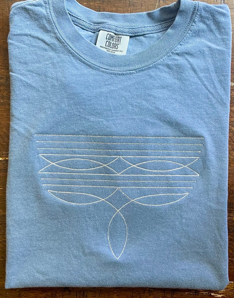 Puede incluir: Una camiseta azul claro con un dise&ntilde;o bordado blanco en la parte delantera. El dise&ntilde;o presenta una serie de l&iacute;neas y formas curvas. La camiseta tiene una etiqueta que dice "Comfort Colors" y "Heavyweight Garment Dyed".