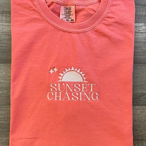 May include: A coral pink t-shirt with a white embroidered design of a sun with two stars above it and the text "SUNSET CHASING". The t-shirt has a tag that reads "COMFORT COLORS" and "XL - TG - EG".