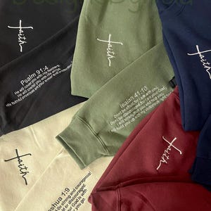 Pode incluir: Uma coleção de camisolas em várias cores, incluindo preto, verde-oliva, azul marinho e bordô. Cada camisola apresenta a palavra "faith" bordada em branco, juntamente com um versículo bíblico na manga. As camisolas são feitas de um material macio.