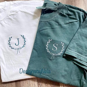 May include: Two t-shirts with embroidered wreaths and initials. The left shirt is white with a 'J' in the wreath. The right shirt is dark green with an 'S' in the wreath. The text 'DesignsbyNela' is visible on the bottom of the shirts.