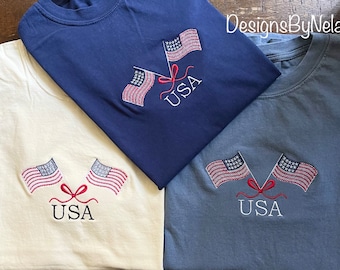 May include: Three t-shirts with embroidered American flags and the word "USA" on them. The shirts are in different colors: navy blue, white, and light blue.