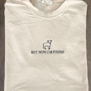 K&ouml;nnte beinhalten: Ein wei&szlig;es T-Shirt mit einem schwarzen gestickten Bild eines Lammes und dem Text "BUT NOW I'M FOUND".