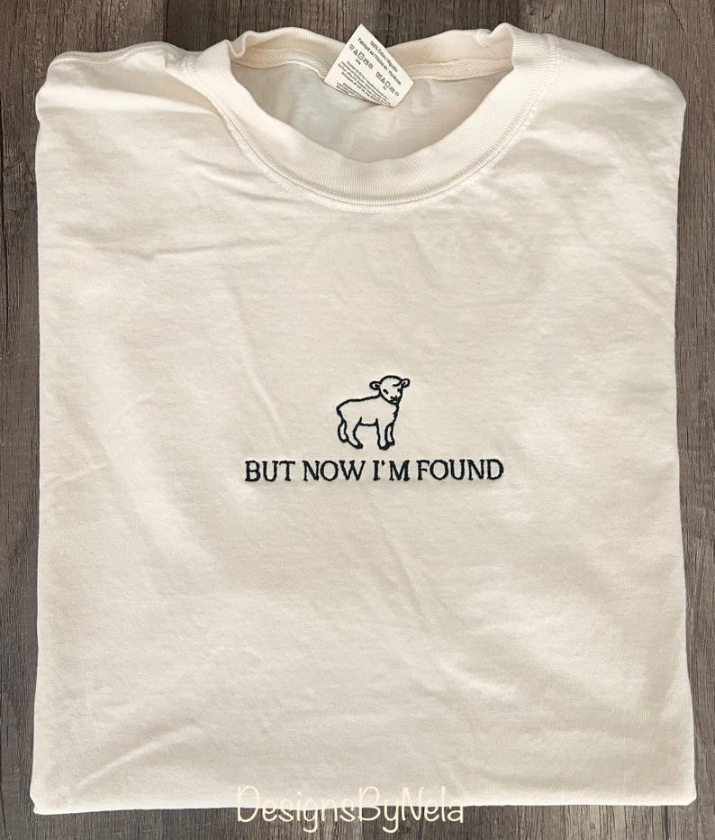 K&ouml;nnte beinhalten: Ein wei&szlig;es T-Shirt mit einem schwarzen gestickten Bild eines Lammes und dem Text "BUT NOW I'M FOUND".