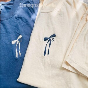 May include: Two short-sleeved t-shirts, one blue and one cream, are shown. Each shirt has an embroidered bow design on the front. The cream shirt has the letter "S" embroidered below the bow. The text "Designs by Nela" is visible at the top.
