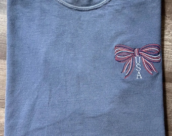 May include: A blue t-shirt with a red, white, and blue bow embroidered on the front. The bow has the word "USA" written in white inside of it.